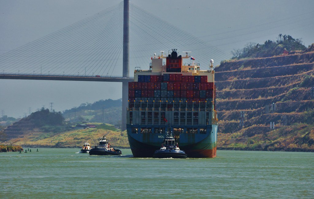 Un gran buque portacontenedores navegando por el Canal de Panamá bajo el Puente de las Américas, asistido por remolcadores.