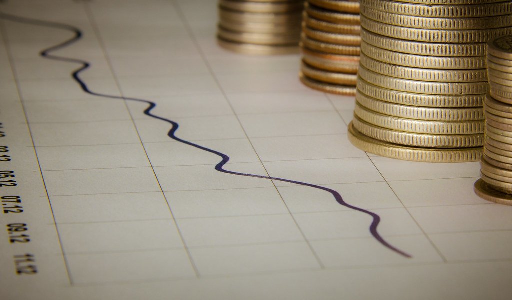 Gráfico de líneas descendente sobre papel cuadriculado junto a varias pilas de monedas doradas.
