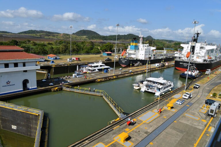 Vista aérea de las esclusas del Canal de Panamá con varios barcos de carga y embarcaciones turísticas transitando por el canal bajo un cielo despejado.
