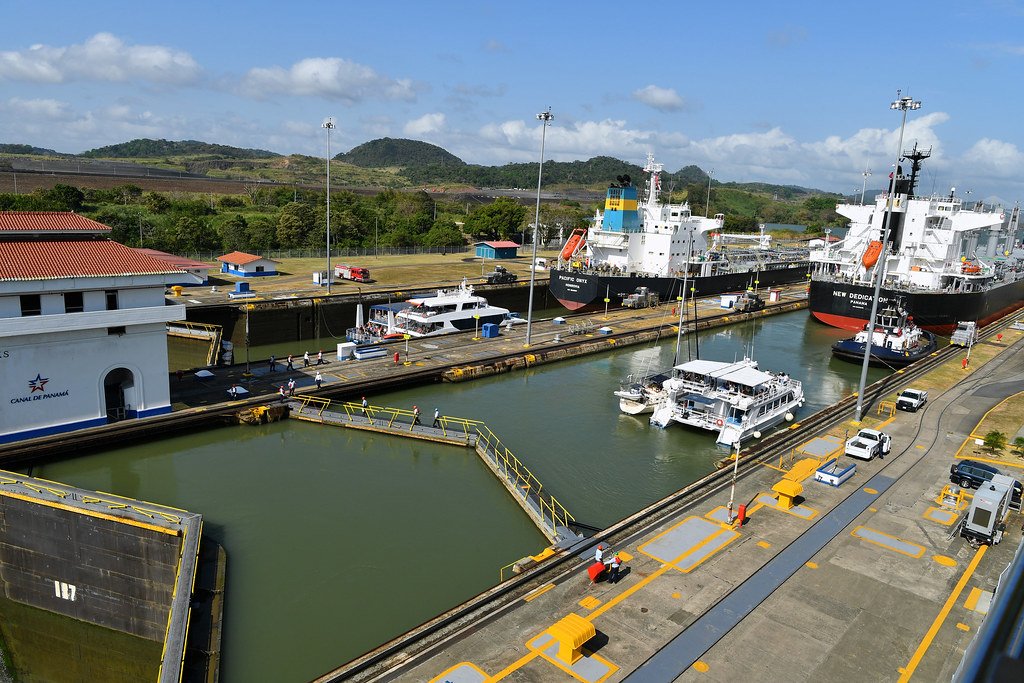Vista aérea de las esclusas del Canal de Panamá con varios barcos de carga y embarcaciones pequeñas transitando, rodeados por instalaciones portuarias y vegetación bajo un cielo despejado.