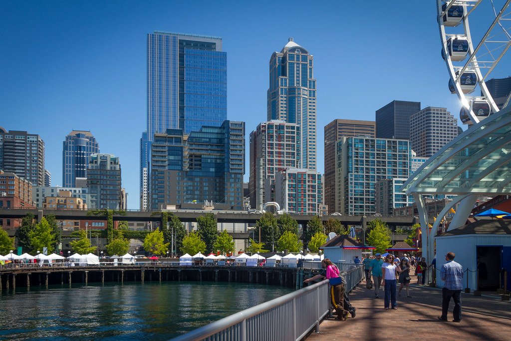 Vista panorámica del centro de Seattle con rascacielos, una noria en primer plano y un muelle con personas caminando bajo un cielo despejado.