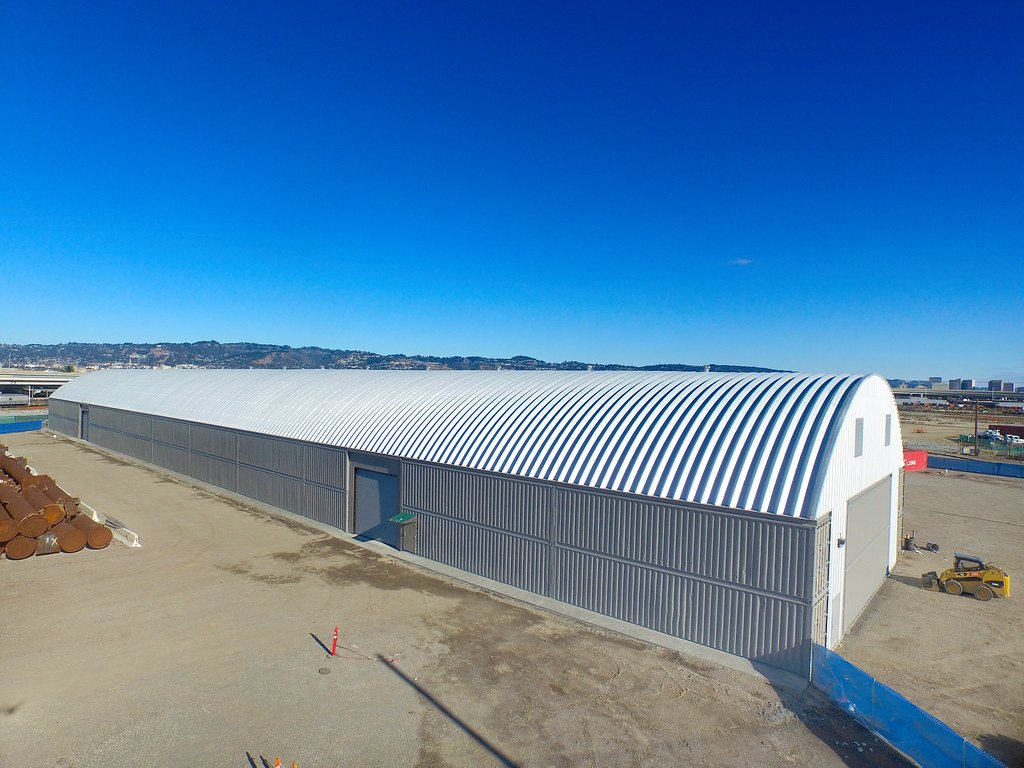 Vista aérea de un almacén industrial de techo arqueado y paredes de metal en un terreno despejado bajo un cielo azul despejado.