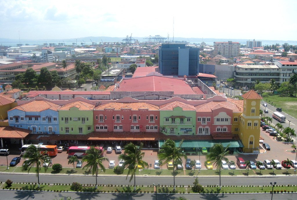 Vista aérea de la Zona Libre de Colón en Panamá, mostrando edificios coloridos de estilo colonial, estacionamientos y una torre, con un puerto y grúas de carga al fondo.