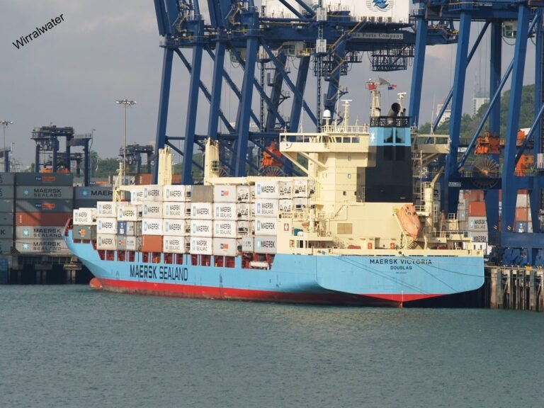 El buque portacontenedores Maersk Victoria atracado en un puerto de carga con grúas pórtico operando en el fondo.
