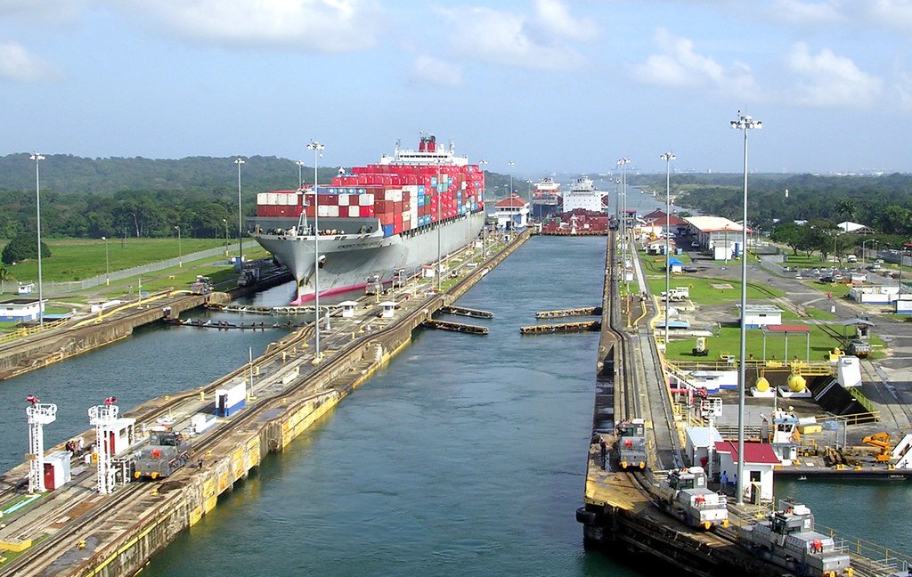 Un gran buque portacontenedores navegando a través de las esclusas del Canal de Panamá, rodeado de vegetación y maquinaria de remolque.