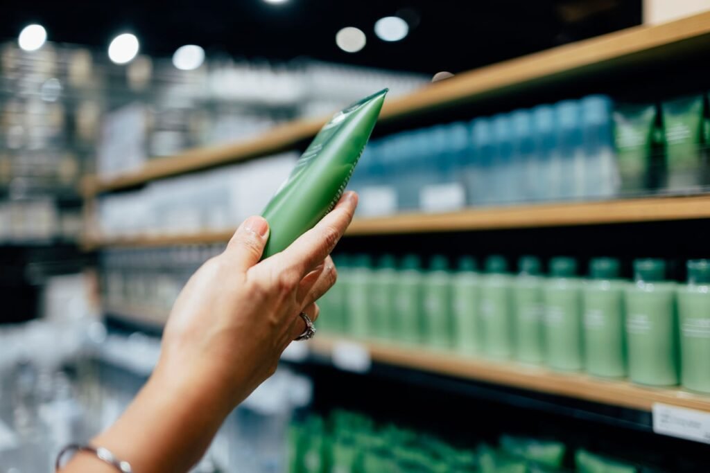 Una mano sostiene un envase verde de cosmético frente a un estante de productos