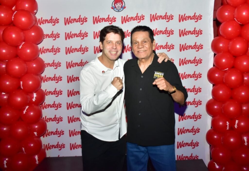 Dos hombres con los puños en alto ante un panel con el logotipo de Wendy’s repetido y columnas de globos rojos a los lados.
