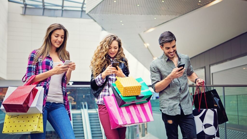 Tres personas jóvenes caminan por la planta alta de un centro comercial con varias bolsas de compras mientras miran sus teléfonos.