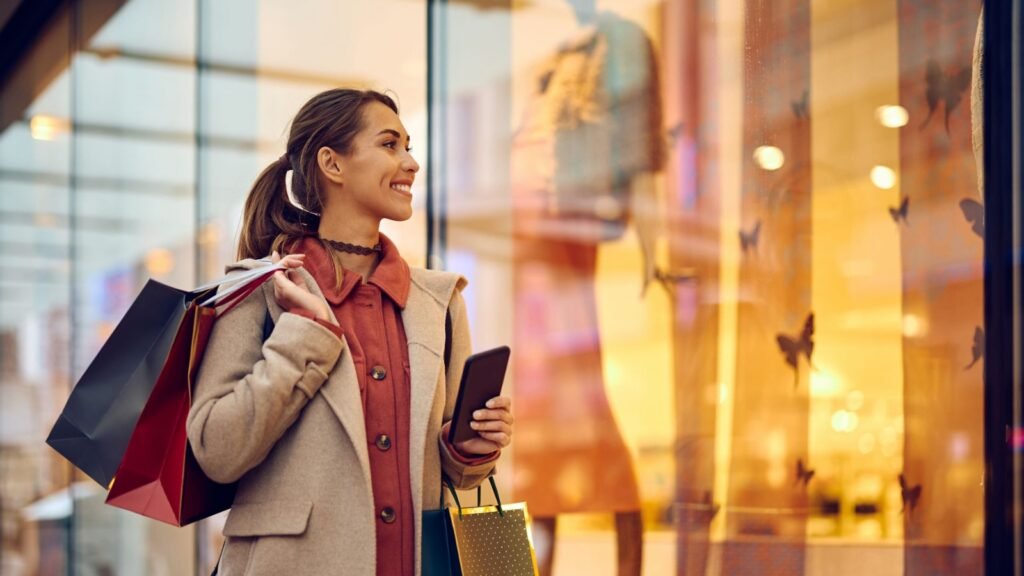 Una mujer con bolsas de compras al hombro y un teléfono en la mano observa el escaparate iluminado de una tienda de ropa.