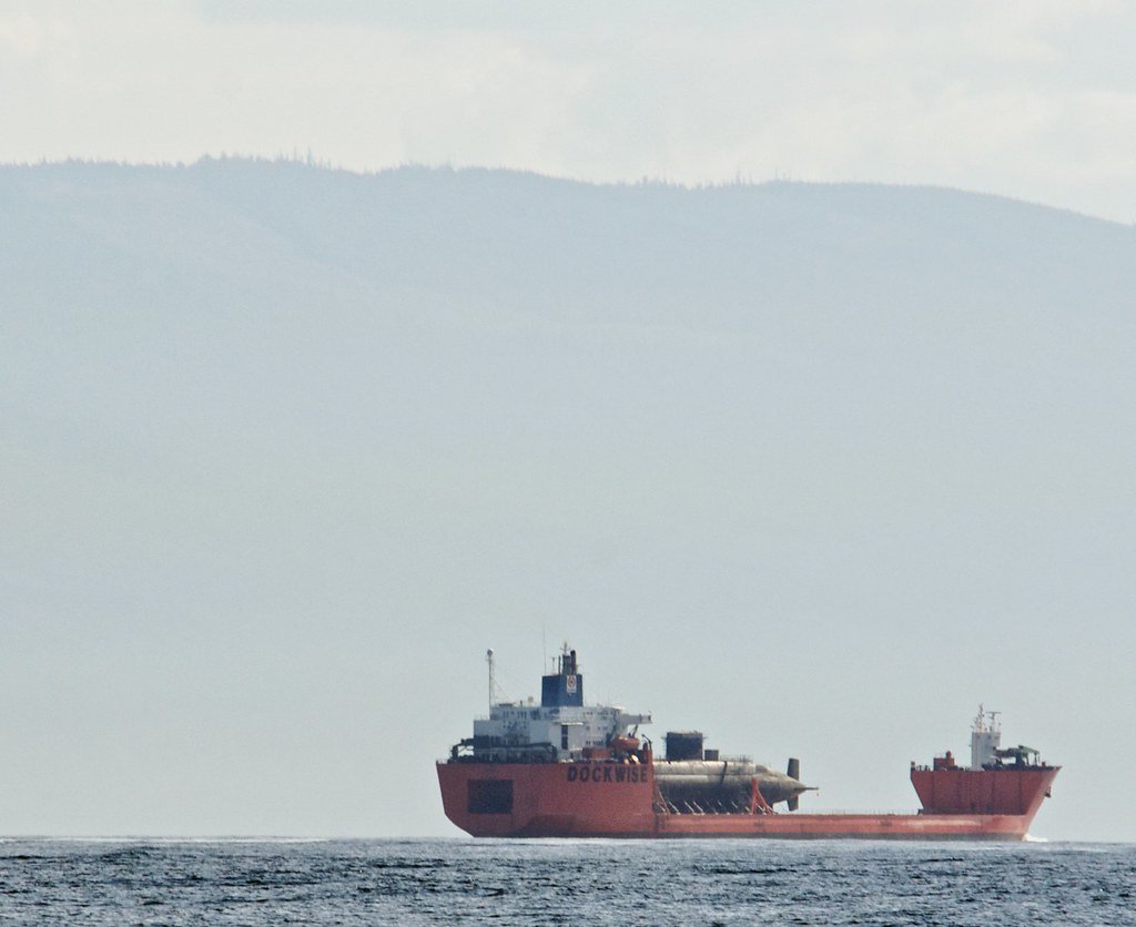 Un buque de carga pesada de la empresa Dockwise transportando un submarino en alta mar con montañas al fondo.