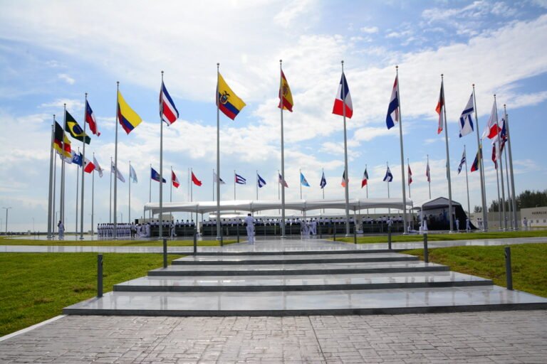 Vista de una explanada con numerosas banderas de diferentes países izadas en mástiles frente a una estructura techada donde se observa personal uniformado en formación.