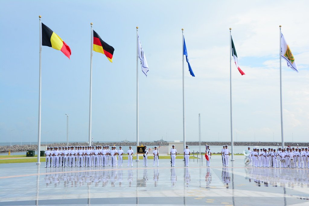 Ceremonia militar con marineros formados frente a seis banderas izadas en mástiles altos cerca de la costa.